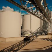Row of large white industrial storage tanks with a metal framework and shadows on a concrete pad under a blue sky.