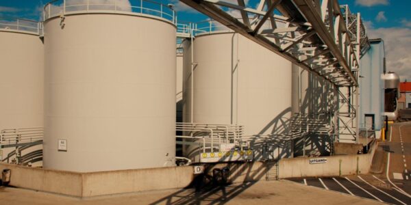 Row of large white industrial storage tanks with a metal framework and shadows on a concrete pad under a blue sky.