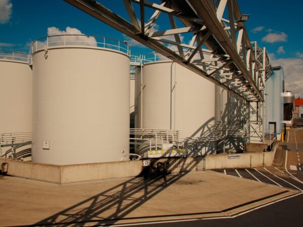 Row of large white industrial storage tanks with a metal framework and shadows on a concrete pad under a blue sky.