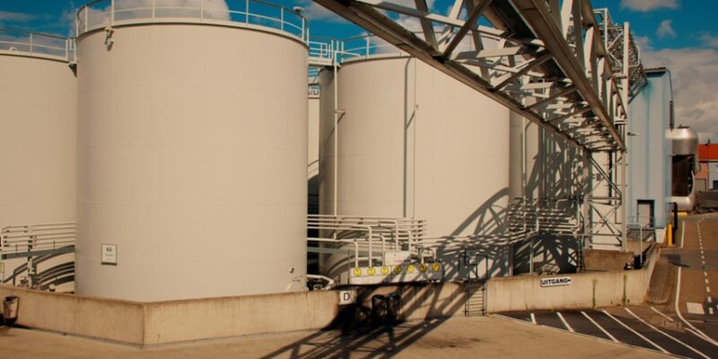 Row of large white industrial storage tanks with a metal framework and shadows on a concrete pad under a blue sky.
