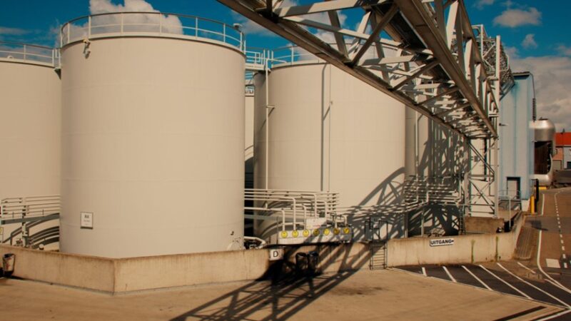 Row of large white industrial storage tanks with a metal framework and shadows on a concrete pad under a blue sky.